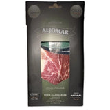 Elegant black packaging of Aljomar Iberian ham. The transparent window in the center reveals bright red, finely marbled ham slices. The Aljomar logo is visible at the top, with "Taste it Love it" and "Product of Spain" mentions on the sides. The packaging highlights the product's "100% Natural" and "A Family Tradition" qualities.