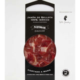 Elegant packaging of Aljomar's 100% Ibérico Bellota Ham. A large circular window reveals finely marbled ham slices with a characteristic deep red color. The Aljomar logo and "Cortado a mano" (hand-sliced) mention are clearly visible, along with a "no lactosa" (lactose-free) label and an expert recommendation, emphasizing the product's premium quality.